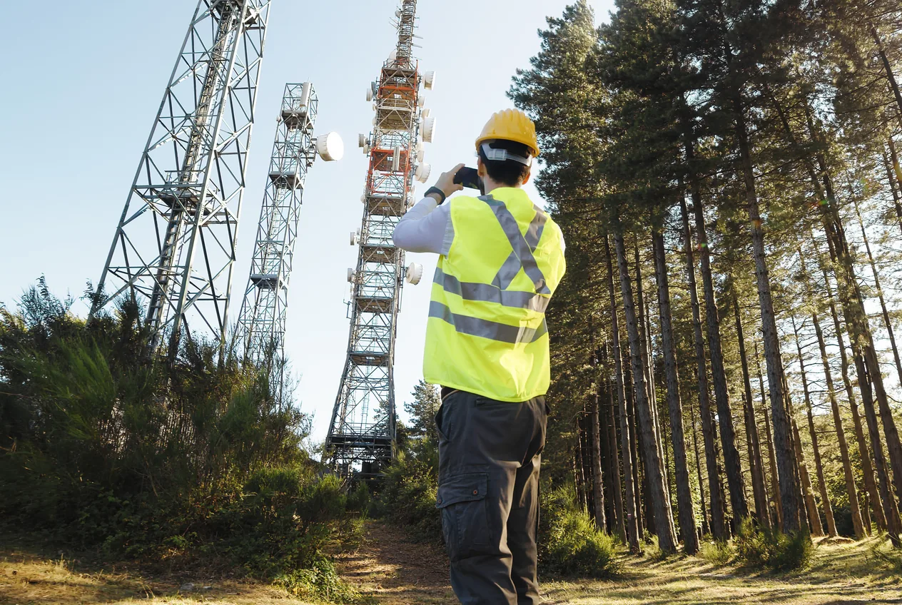 A worker in a safety vest photographing telecommunications towers at the edge of a forest